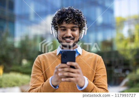 Portrait of smiling man wearing headphones, using smartphone outdoors in urban environment. Person enjoying music with phone. Capturing moment of happiness and connectivity, highlighting technology Portrait of smiling man wearing headphones, using smartphone outdoors in urban environment. Person enjoying music with phone. Capturing moment of happiness and connectivity, highlighting technology 117993727