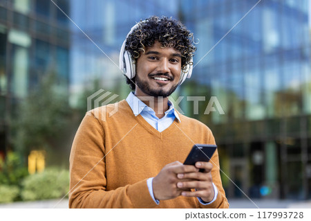 Confident young man with curly hair wearing headphones and holding smartphone outside contemporary glass office building. He looks happy and relaxed, dressed in casual sweater Confident young man with curly hair wearing headphones and holding smartphone outside contemporary glass office building. He looks happy and relaxed, dressed in casual sweater 117993728