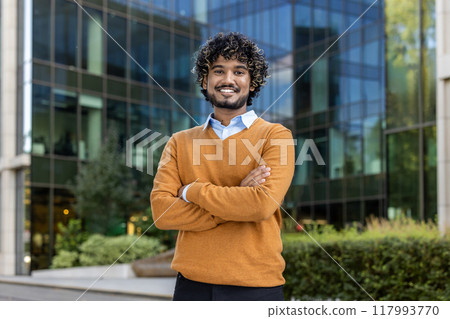 Confident businessman stands outside modern office building, wearing stylish orange sweater. His expression exudes success and professionalism, embodying urban business environment's dynamic energy. Confident businessman stands outside modern office building, wearing stylish orange sweater. His expression exudes success and professionalism, embodying urban business environment's dynamic energy. 117993770
