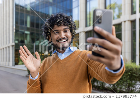 Cheerful man taking selfie with phone in city environment, wearing casual clothes. Captures joyful expression, waving to camera, experiencing happiness and connection. Ideal for themes of technology 117993774