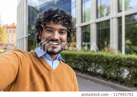 Man with curly hair taking selfie in urban area. Wearing orange sweater over blue shirt. Smiling broadly, enjoying outdoor setting. Technology emphasizes friendly and approachable nature. Man with curly hair taking selfie in urban area. Wearing orange sweater over blue shirt. Smiling broadly, enjoying outdoor setting. Technology emphasizes friendly and approachable nature. 117993781
