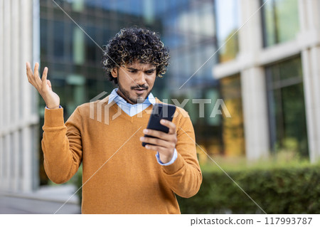 Man with curly hair standing outside modern office building looks at phone with frustration. Wearing a brown sweater, he expresses confusion or dissatisfaction with technology issues. Man with curly hair standing outside modern office building looks at phone with frustration. Wearing a brown sweater, he expresses confusion or dissatisfaction with technology issues. 117993787