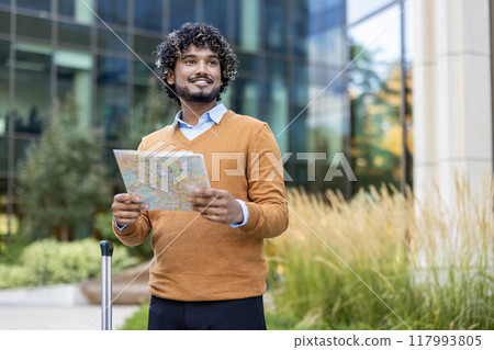 Confident man stands outdoors holding map, looking away with a smile. Dressed in casual attire, he exudes positivity. Urban setting in background suggests travel, exploration. Perfect for travel 117993805