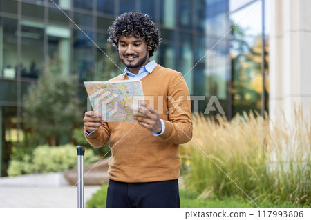 Man holding a map in modern city setting for urban exploration. He appears focused and prepared for travel or journey, with suitcase beside him. Autumn vibes with greenery and modern architecture. 117993806