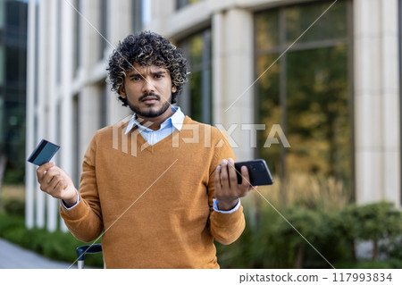 Hispanic businessman stands outdoors holding credit card and phone, expressing concern outside modern office building. Scene captures financial or business challenge 117993834