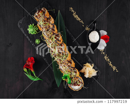 Tempura maki sushi roll lying on black slate plate decorated with flowers Tempura maki sushi roll lying on black slate plate decorated with flowers 117997219