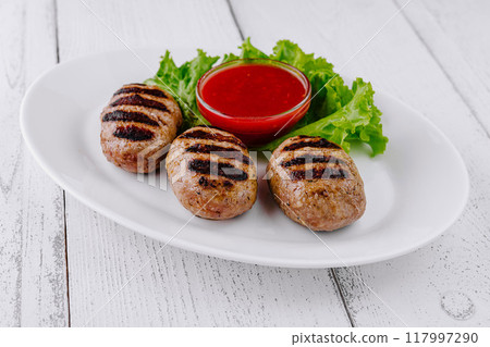 Three grilled meatballs with red sauce on white plate resting on white table 117997290