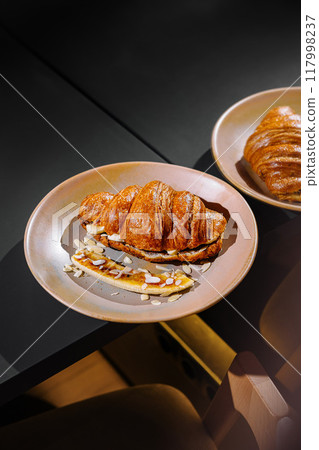 Delicious croissants with banana and almonds on plates on a table Delicious croissants with banana and almonds on plates on a table 117998237