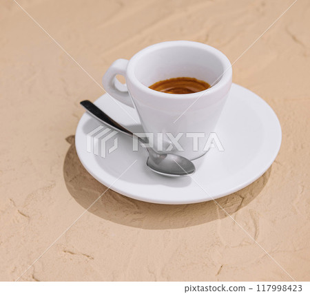 A white cup of espresso coffee on a sandy surface under soft sunlight 117998423