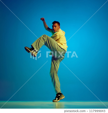 Energy of hip-hop. Artistic and creative young man, dancer performing with passion against gradient blue background in neon light Energy of hip-hop. Artistic and creative young man, dancer performing with passion against gradient blue background in neon light 117999843