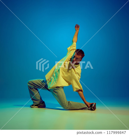 Energy of hip-hop. Artistic and creative young man, dancer performing with passion against gradient blue background in neon light 117999847