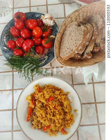 A plate with oriental pilaf on the table near a plate with bread and tomatoes. Concept of delicious home cooking in nature. A plate with oriental pilaf on the table near a plate with bread and tomatoes. Concept of delicious home cooking in nature. 118000593