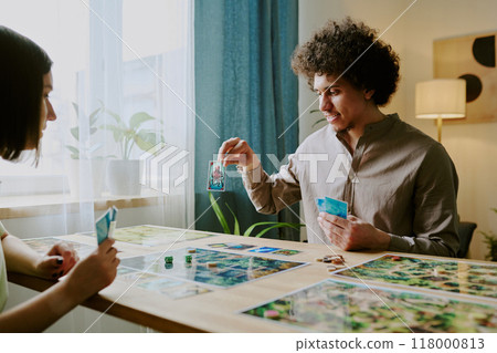 Young biracial man showing his card to girlfriend while playing modern fantasy board game together at home 118000813