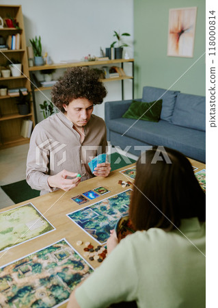 Vertical high angle view of young biracial man sitting at table in living room taking dice while playing board game with his girlfriend Vertical high angle view of young biracial man sitting at table in living room taking dice while playing board game with his girlfriend 118000814