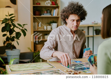 Young biracial man with curly hair sitting at table in living room and playing board game with unrecognizable girl friend Young biracial man with curly hair sitting at table in living room and playing board game with unrecognizable girl friend 118000815