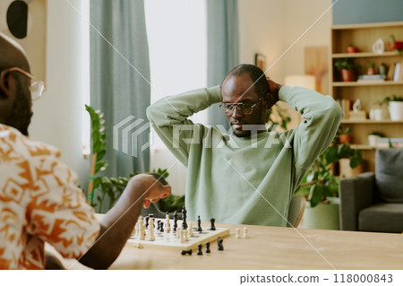 Young Black man sitting at wooden table with hands behind his head watching friend moving chess piece during game 118000843