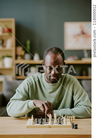 Vertical shot of young African American man sitting at wooden desk in living room solving chess problem, copy space Vertical shot of young African American man sitting at wooden desk in living room solving chess problem, copy space 118000912