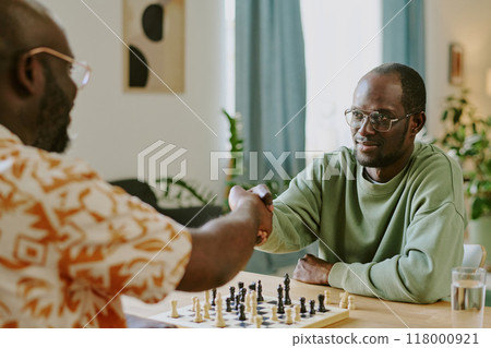 Over shoulder shot of two young African American men doing handshake while playing chess game in living room 118000921