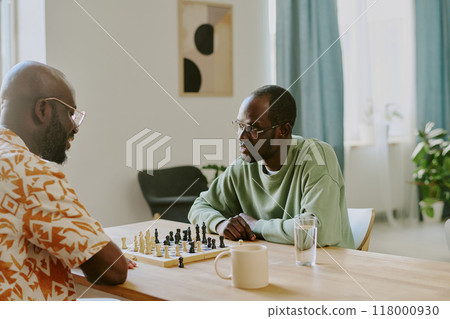 Two young African American male friends sitting at table in front each other and talking while playing chess in living room 118000930
