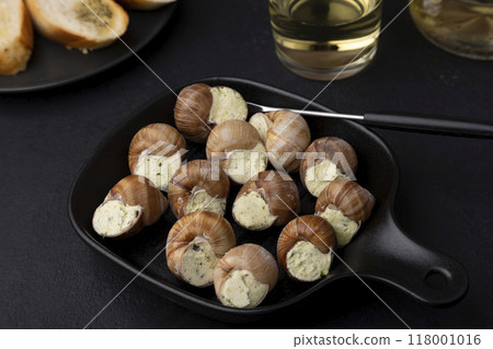 Escargot bourguignon with butter and garlic. Traditional French dish made from snails, served with baguette and white wine. Selective focus, close-up, black background. 118001016