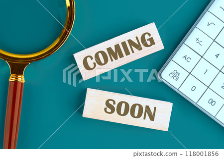 Coming soon text written it is laid out from wooden blocks , top view in a composition with a calculator and a magnifying glass Coming soon text written it is laid out from wooden blocks , top view in a composition with a calculator and a magnifying glass 118001856