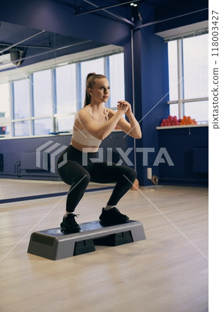 Woman performing squats on a step platform in a bright fitness studio 118003427