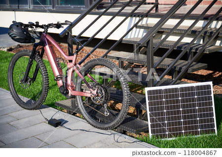 Pink electric mountain bike parked beside staircase, connected to small solar panel for charging. Bike rugged tires, mounted helmet on handlebar, situated on paved pathway with grassy lawn. 118004867
