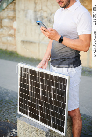 Cropped photo of man charging phone by portable solar panel. Young male using solar battery charger for mobile devices. Concept of alternative energy resources and sustainable lifestyle. Cropped photo of man charging phone by portable solar panel. Young male using solar battery charger for mobile devices. Concept of alternative energy resources and sustainable lifestyle. 118004883