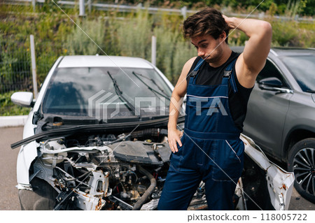 Confused mechanic scratching head in frustration while assessing damage on car after an accident. Vehicle service repairman looking car front end completely destroyed in garage car service. Confused mechanic scratching head in frustration while assessing damage on car after an accident. Vehicle service repairman looking car front end completely destroyed in garage car service. 118005722