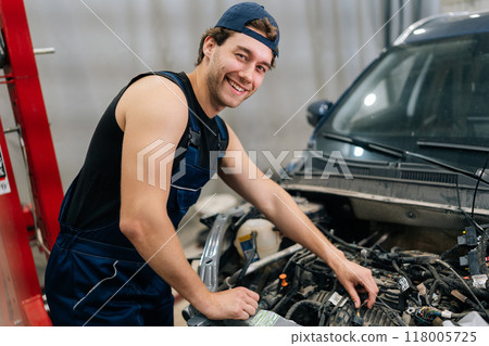 Portrait of skilled young mechanic joyfully examining car engine in auto repair shop smiling looking at camera. Cheerful repairman happily inspecting vehicle engine in car service. Portrait of skilled young mechanic joyfully examining car engine in auto repair shop smiling looking at camera. Cheerful repairman happily inspecting vehicle engine in car service. 118005725