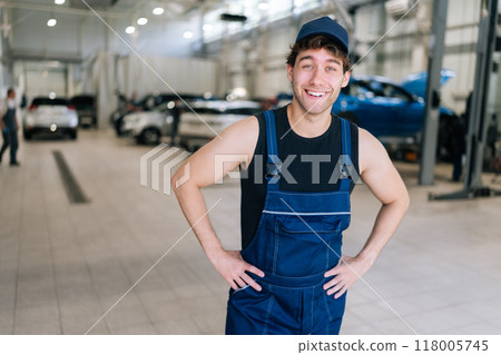 Portrait of joyful repairman in uniform standing smiling looking at camera in auto repair shop indoors. Confident professional talented mechanic male posing to camera, ready to repair vehicle. 118005745