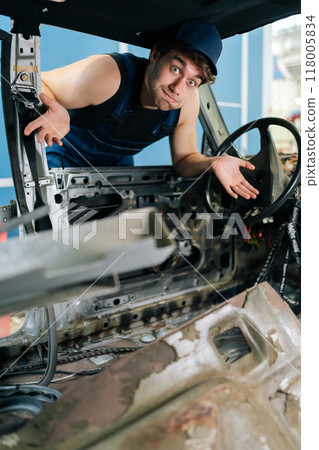 Vertical portrait of puzzled mechanic male gesturing inside disassembled car, showcasing automotive expertise and work in progress, looking at camera with puzzled emotion. Shooting in slow motion. 118005834