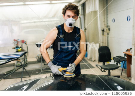 Concept of car detailing and polishing. Portrait of professional car service male worker polishing black luxury car hood in auto repair shop using with orbital polisher, looking at camera. Concept of car detailing and polishing. Portrait of professional car service male worker polishing black luxury car hood in auto repair shop using with orbital polisher, looking at camera. 118005926