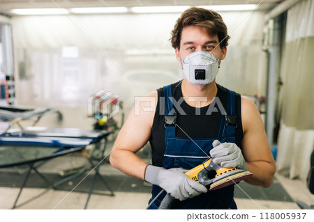 Mechanic male in protective mask holding machinery car polishers maintenance to remove marks repair. Portrait of car paint technician in uniform holding car polisher to polish of car with refinement. 118005937