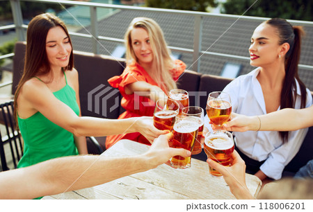Joyful group enjoys cold beers on terrace, clinking glasses and smiling, celebrating friendship and simple pleasures of life. 118006201