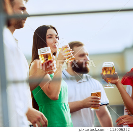 Group of friends enjoys sunny day together, sipping cold beers. Woman in green dress takes drink, surrounded by smiling faces and relaxed vibe. 118006252