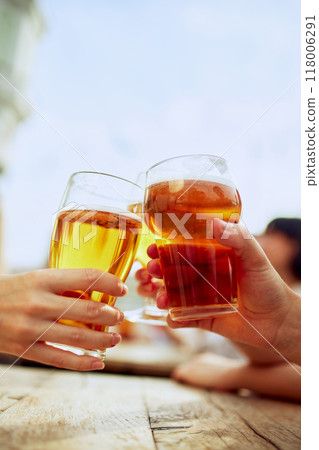Two glasses of beer meet in cheerful toast, capturing essence of friendship and good times. Sunlight reflects in golden liquid, enhancing moment. 118006291