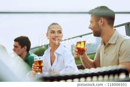 Young bearded man enjoys cold beer in relaxed outdoor setting, savoring moment. Woman sits nearby. Festive atmosphere. 118006301