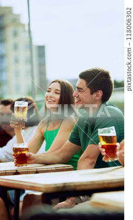 Young bearded man enjoys cold beer in relaxed outdoor setting, savoring moment. Woman sits nearby. Festive atmosphere. 118006302