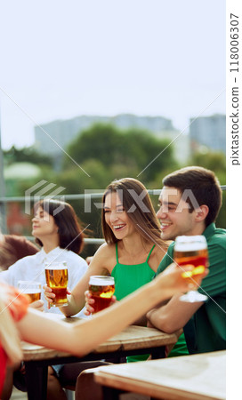Young people sitting on terrace of urban pub, and clinking their beer mugs, celebrating meeting after long years. Selective focus. 118006307