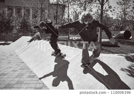 Two riders, active young boys in stylish clothes training outdoors, skateboarding in public park. Black and white photography. Two riders, active young boys in stylish clothes training outdoors, skateboarding in public park. Black and white photography. 118006396