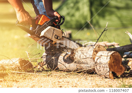 Chain saw in hands of man cutting large tree trunks outdoors while camping. Chain saw in hands of man cutting large tree trunks outdoors while camping. 118007407