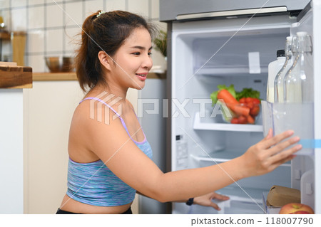 Smiling young woman dressed in casual workout attire reaching for something inside the fridge, preparing for healthy meal Smiling young woman dressed in casual workout attire reaching for something inside the fridge, preparing for healthy meal 118007790