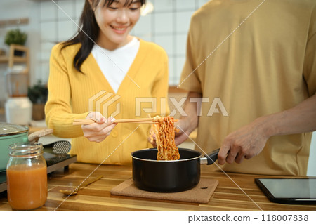 Happy young couple holding a pot with freshly cooked instant noodles, enjoying a casual meal together in kitchen. Happy young couple holding a pot with freshly cooked instant noodles, enjoying a casual meal together in kitchen. 118007838