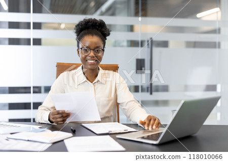 African American businesswoman confidently working at desk in modern office setting. She holds documents and uses laptop, showcasing professionalism and focus. 118010066