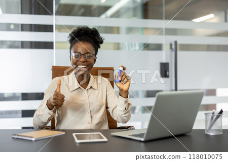 African American businesswoman in office smiling with confidence, holding inhaler and giving thumbs up. Surroundings include laptop, tablet, and phone, creating modern professional setting. 118010075