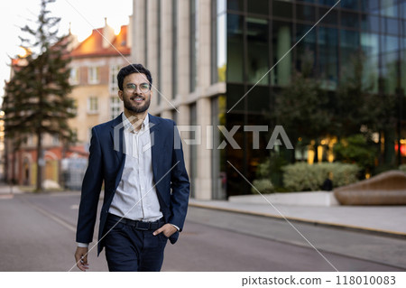 Confident young businessman wearing glasses walking outdoors in city. Smiling professional in jacket and shirt enjoying urban environment. Modern office building in background. Confident young businessman wearing glasses walking outdoors in city. Smiling professional in jacket and shirt enjoying urban environment. Modern office building in background. 118010083