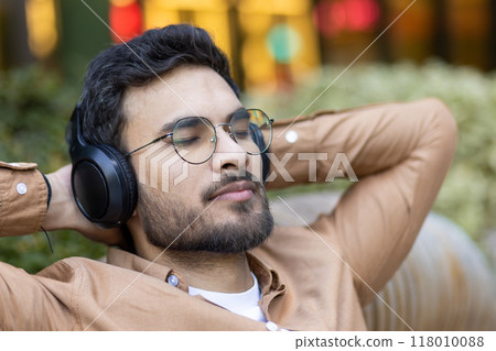Relaxed young man with beard wearing headphones, eyes closed, enjoying music outdoors. Casual attire signifies tranquil break in busy urban park. Relaxed young man with beard wearing headphones, eyes closed, enjoying music outdoors. Casual attire signifies tranquil break in busy urban park. 118010088