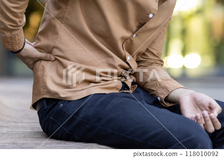 Individual seated on bench outdoors showing signs of discomfort due to lower back pain. Casual attire, hand on back indicating ache or injury. Expresses physical discomfort, health concern 118010092