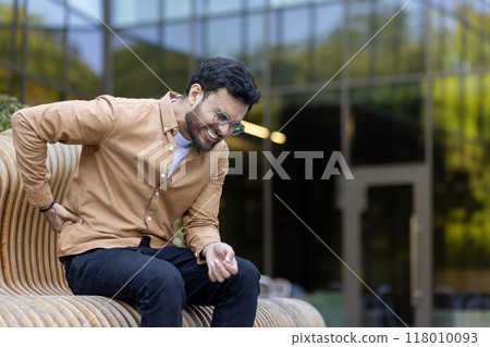 Man seated on outdoor bench holding back in pain, expressing discomfort and stress. Wearing casual brown shirt and glasses, situated in urban environment. Concept of health issues, stress, back pain 118010093